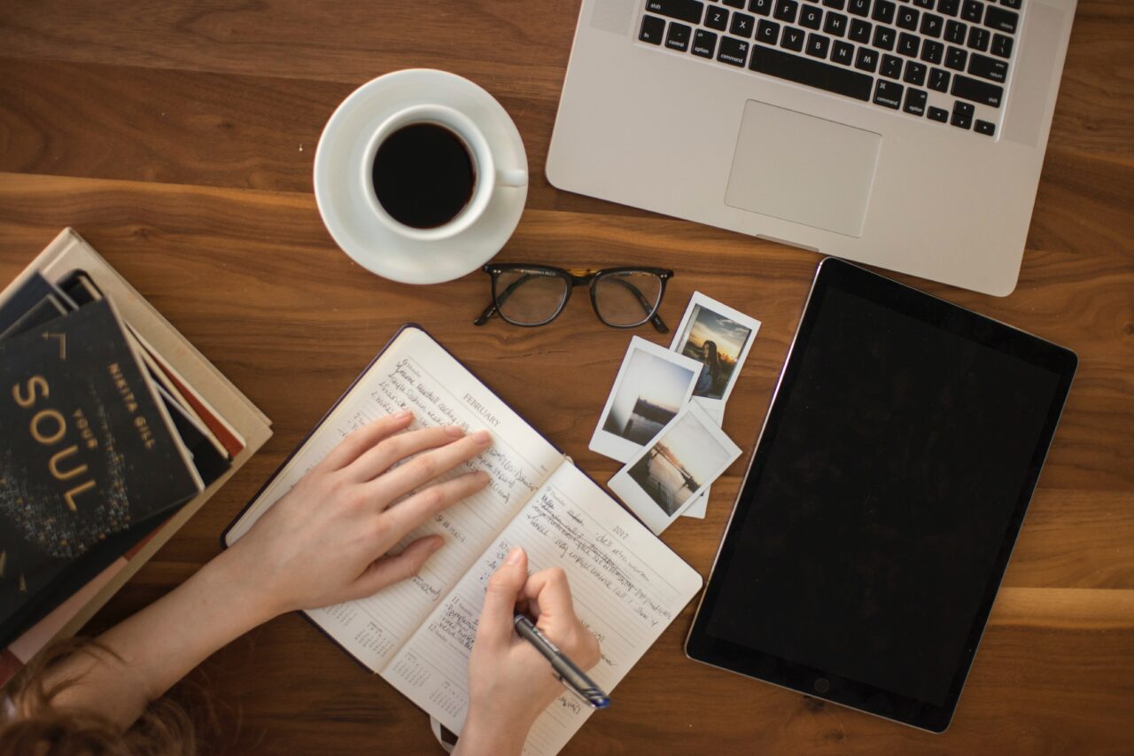 Image of a hand writing notes or brainstorming in a notebook on a wooden table with a cup of coffee, glasses and computer