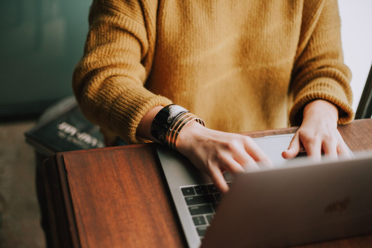 A person's torso in an ochre sweater seated at a laptop with hands poised over the keyboard